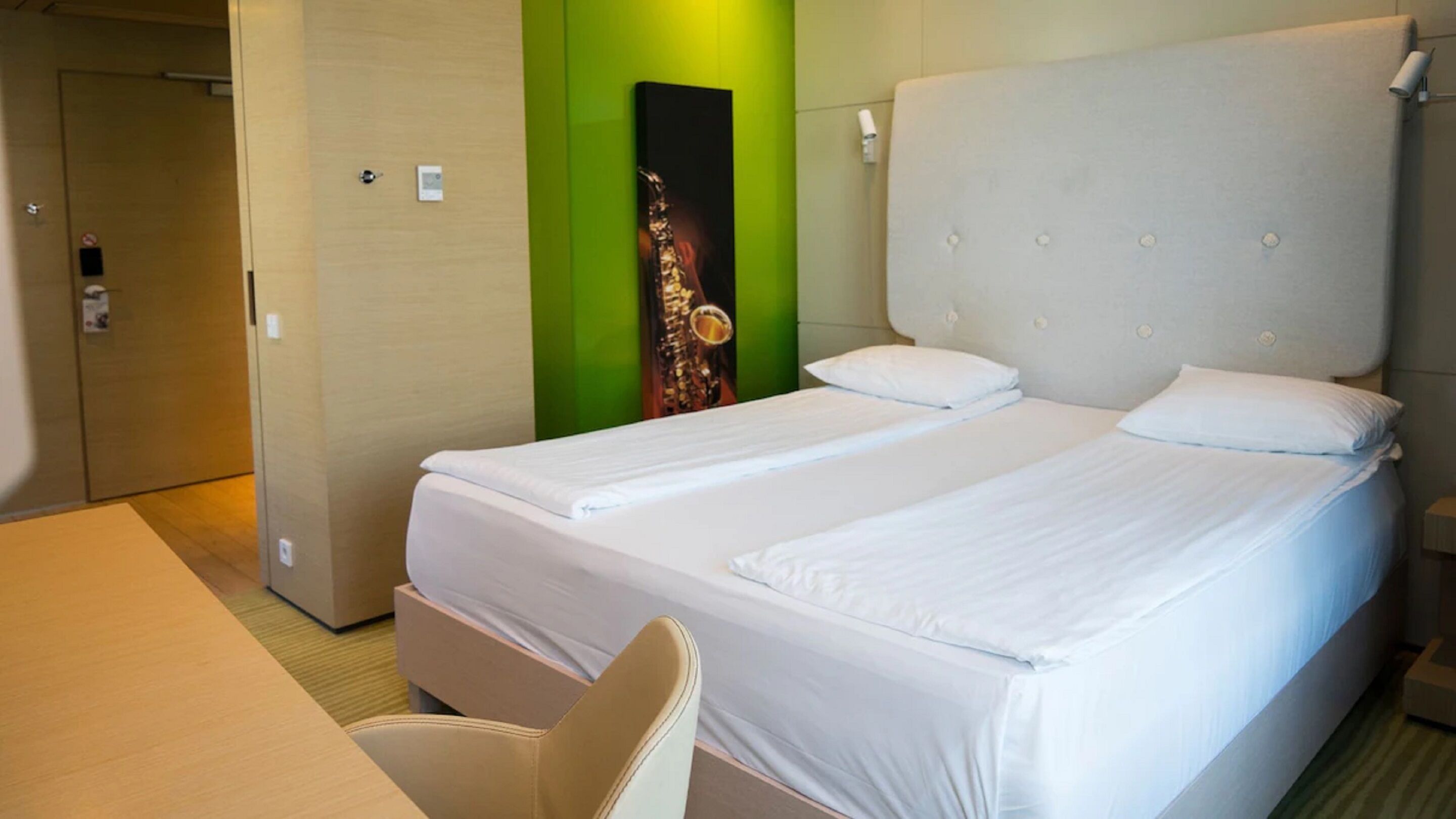 A hotel room with a double bed, a chair, a desk, and a green accent wall with a saxophone poster.
