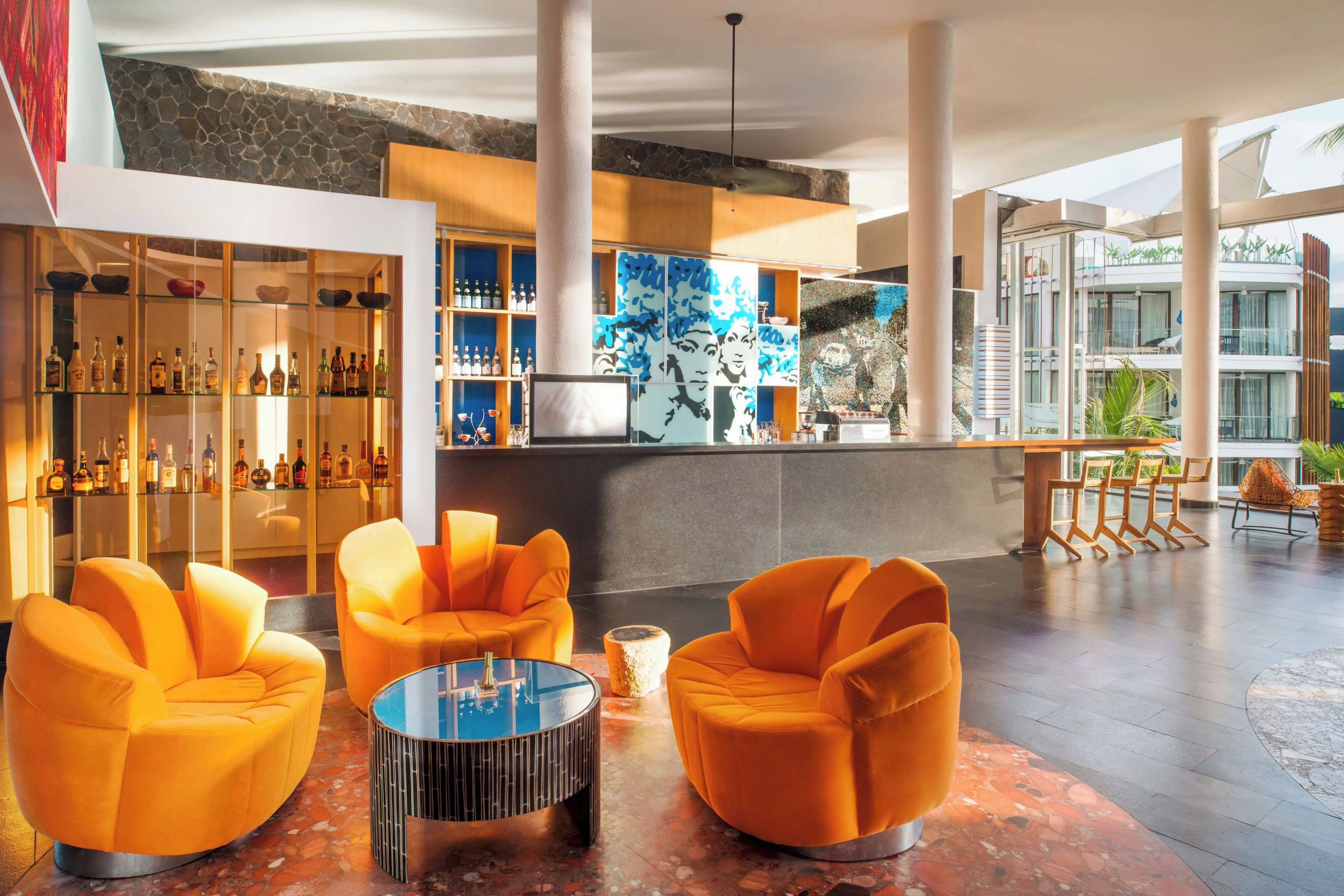 Zona de bar moderna con una vitrina de botellas, asientos color naranja y un amplio ventanal con vistas a un edificio.
