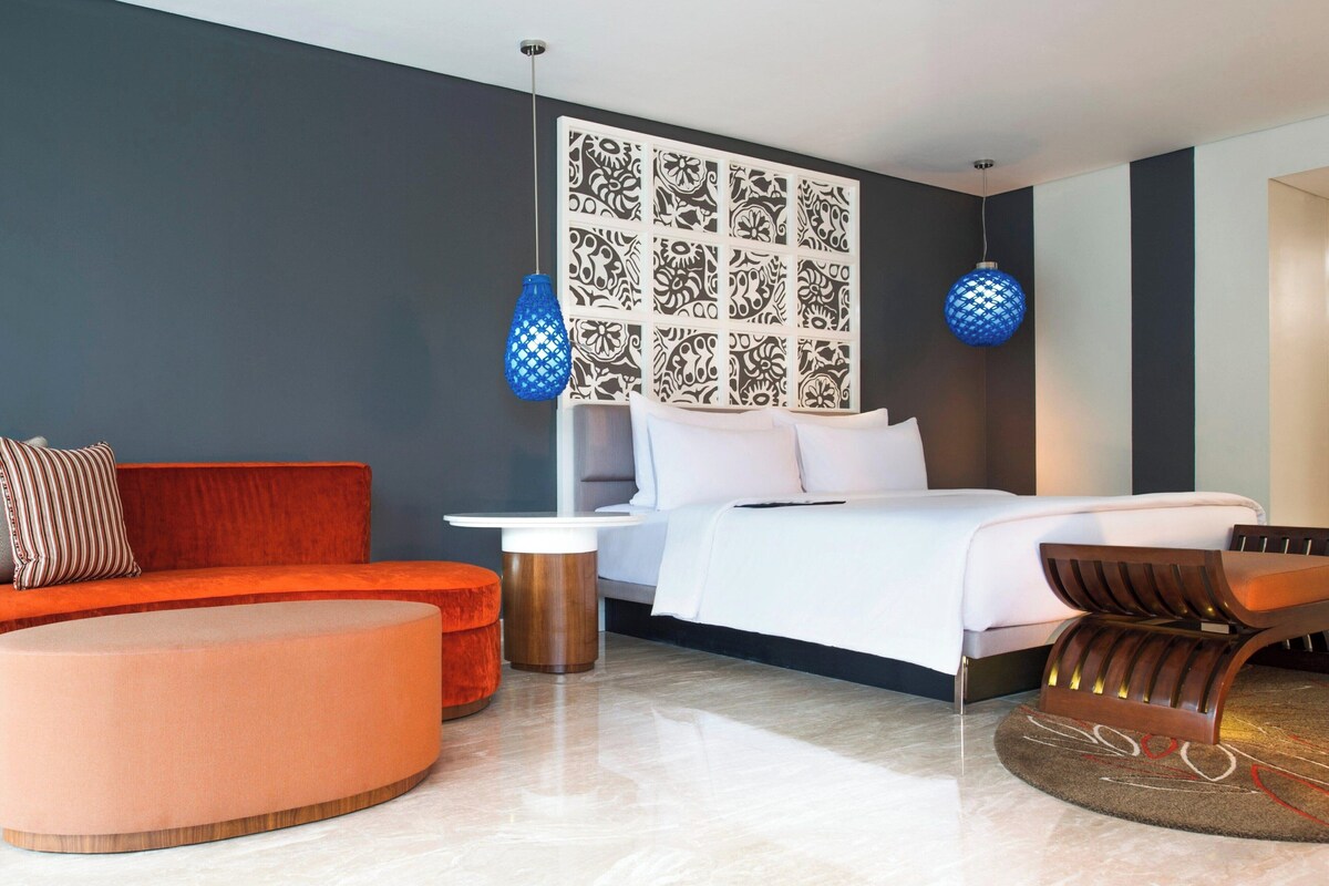 A modern hotel room with a bed, a sofa, a round ottoman, a nightstand, and two wall-mounted lamps.
