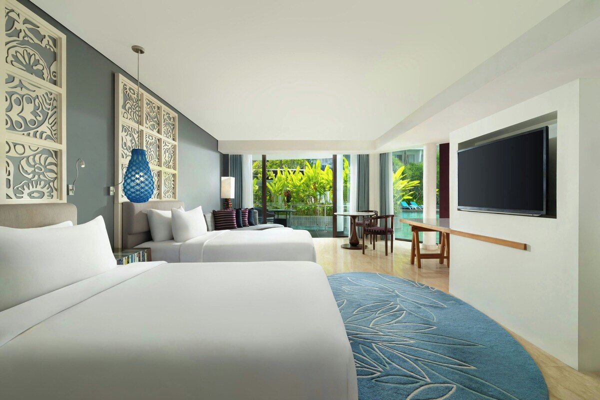 A hotel room with two beds, a TV, a desk, and a balcony with a view of greenery.