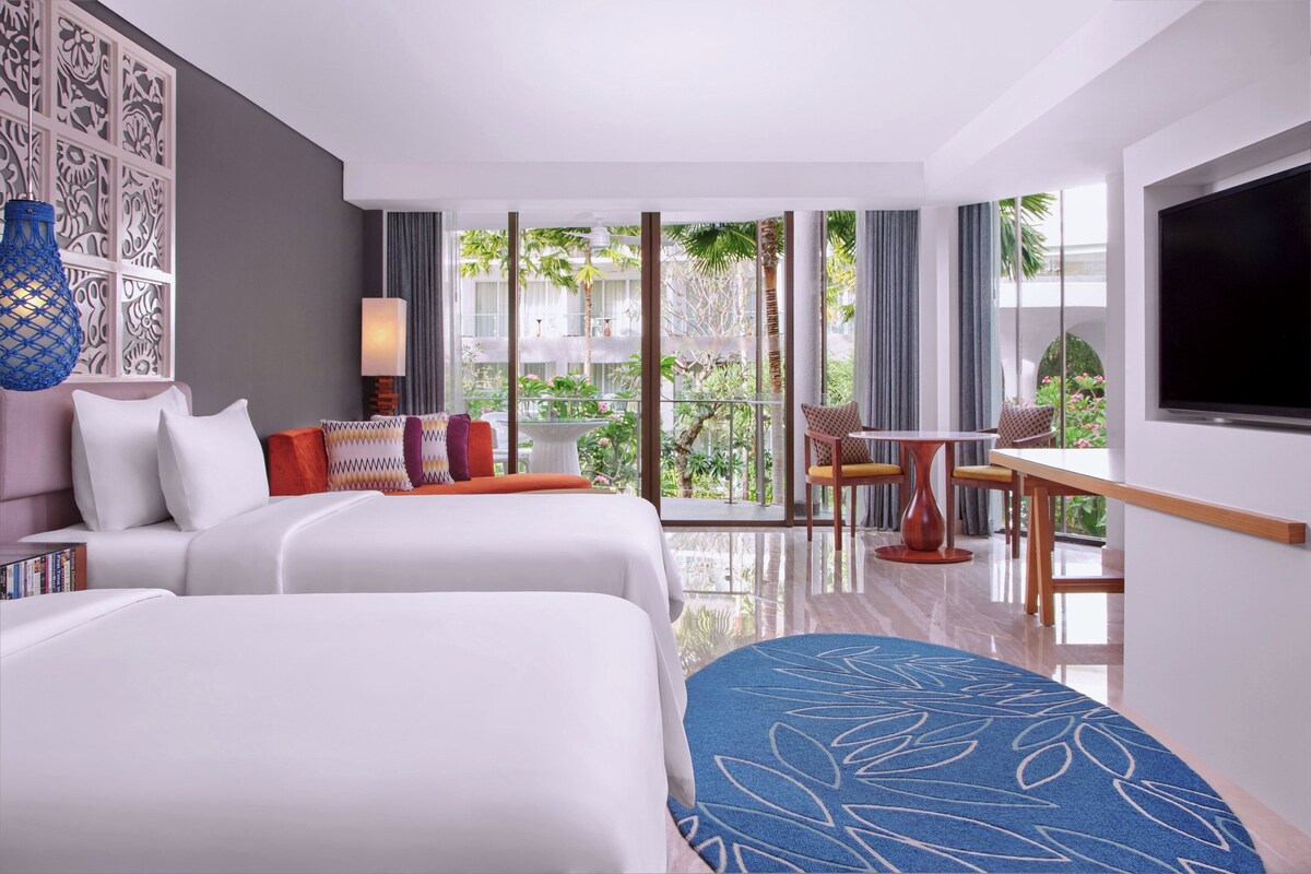 A hotel room with two beds, a dining area, a TV, and a balcony with plants.