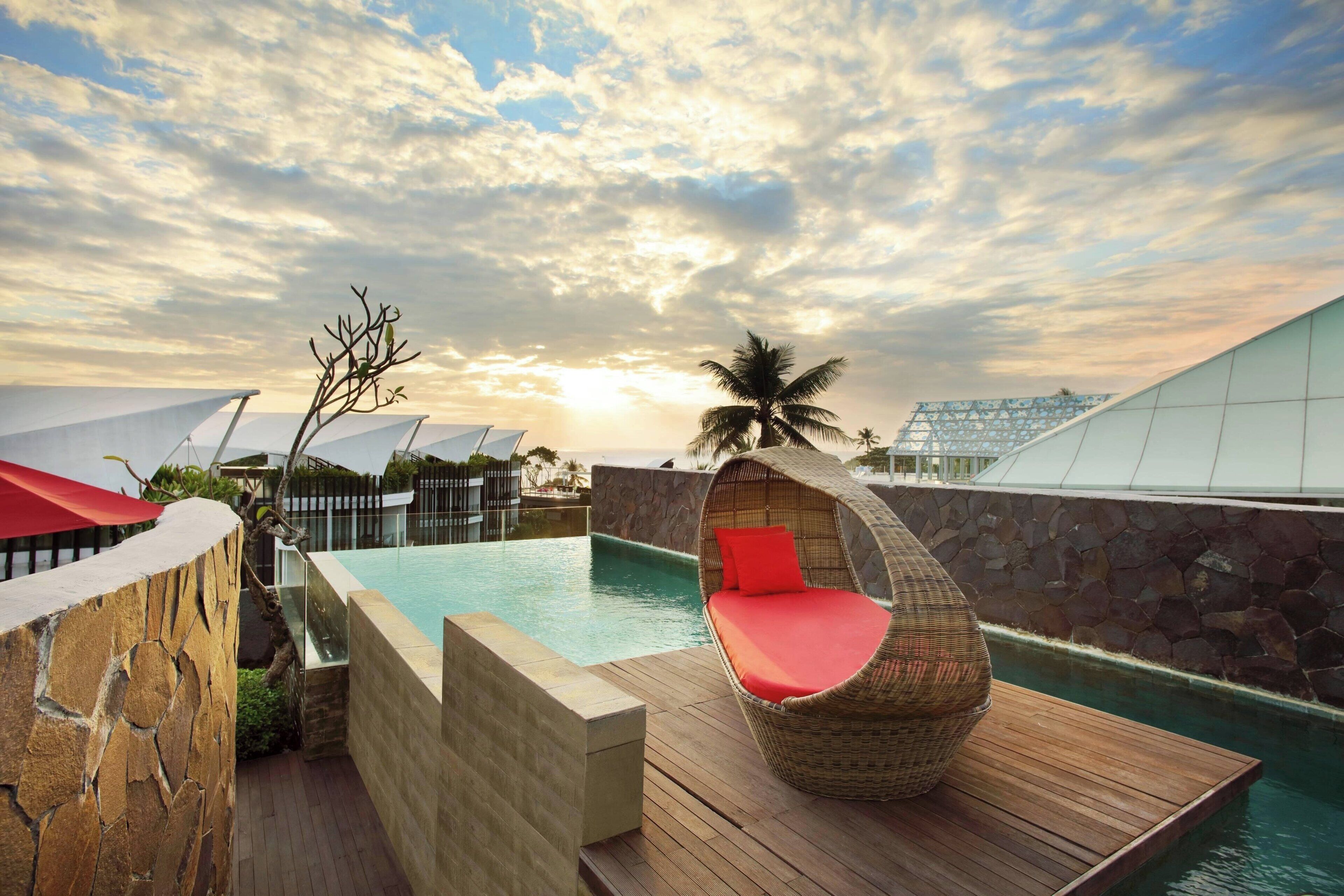 Un área de piscina con cubierta de madera, una silla de mimbre con cojines rojos y una palmera.