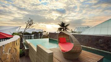 Un área de piscina con cubierta de madera, una silla de mimbre con cojines rojos y una palmera.