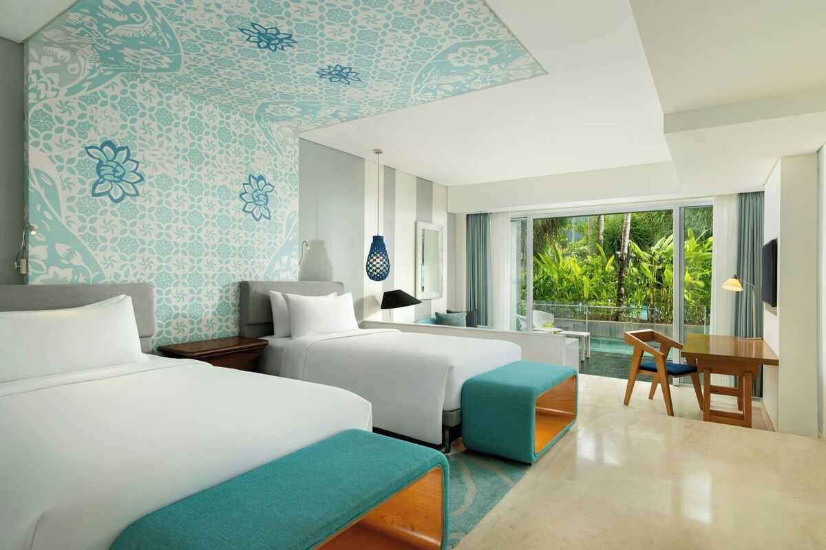 A hotel room with two beds, a desk, and a view of greenery outside.