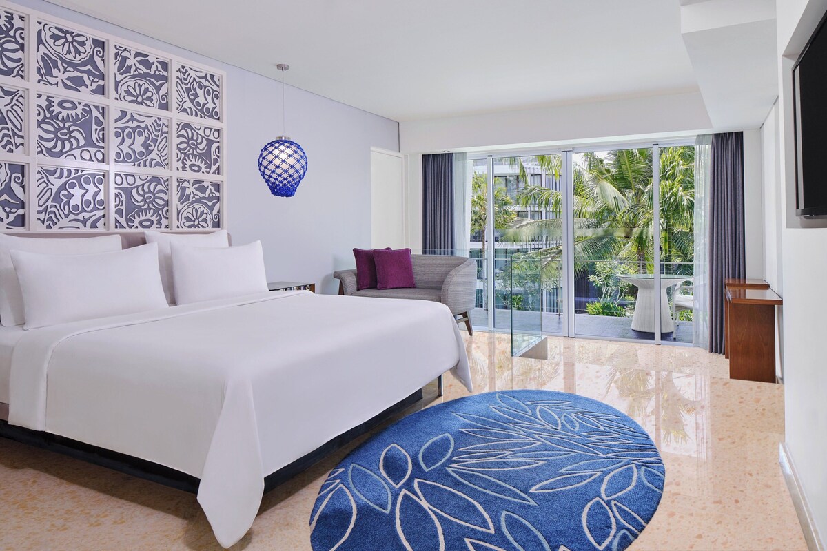 A hotel room with a large bed, a blue patterned rug, a wall with decorative panels, and a view of palm trees through the balcony.