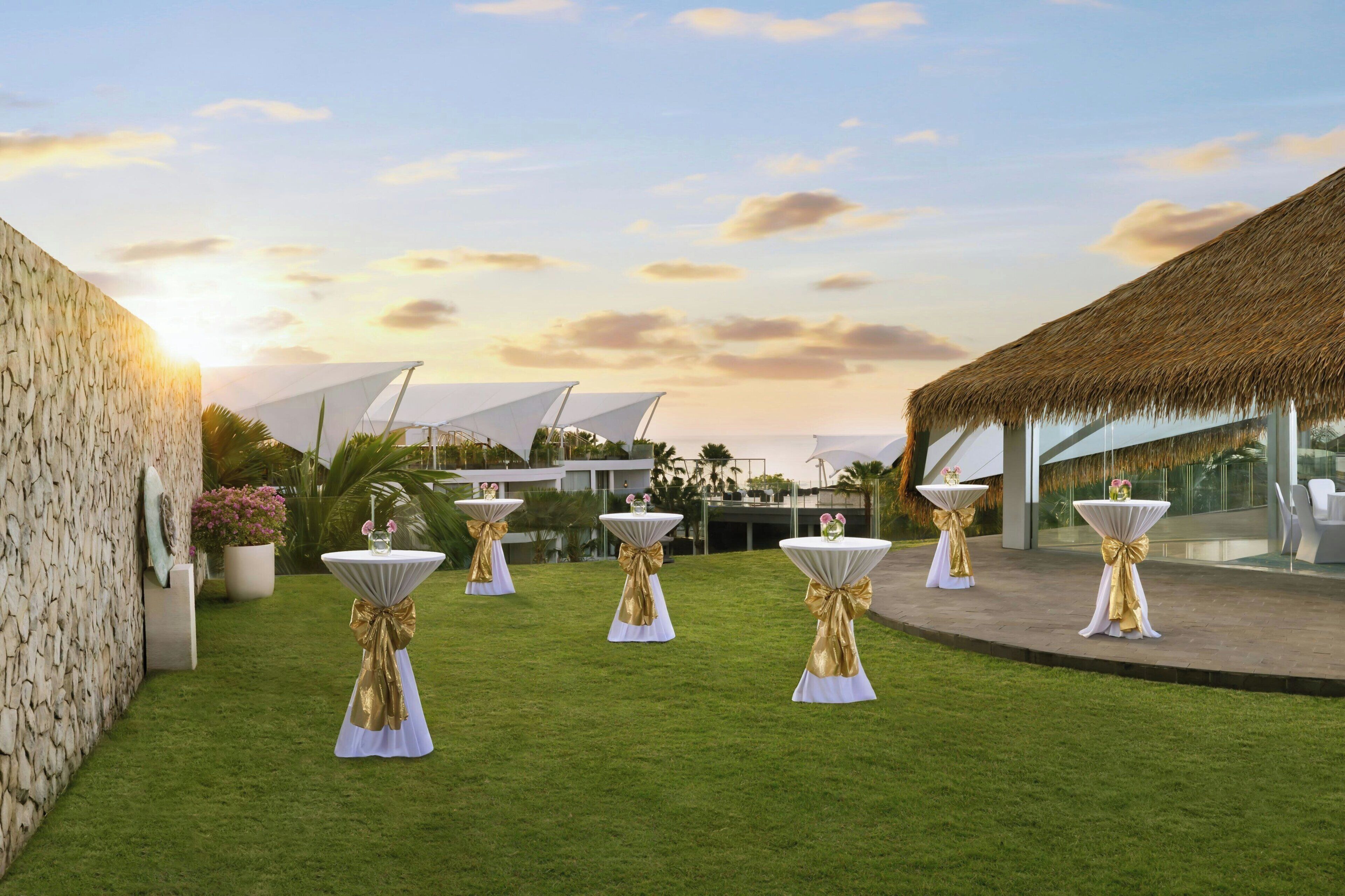 A courtyard with white tables and gold sashes, a stone wall, and a thatched-roof structure.