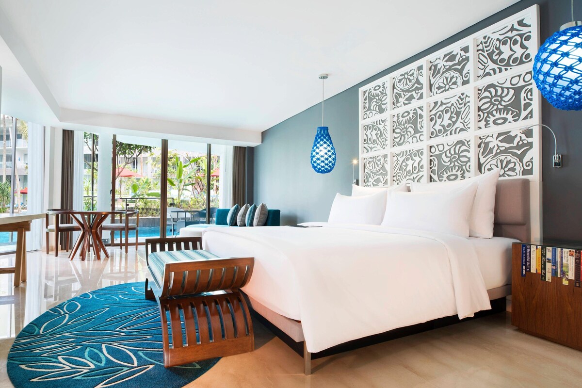 A modern hotel room with a large bed, a dining area with a pool view, and a decorative wall panel.
