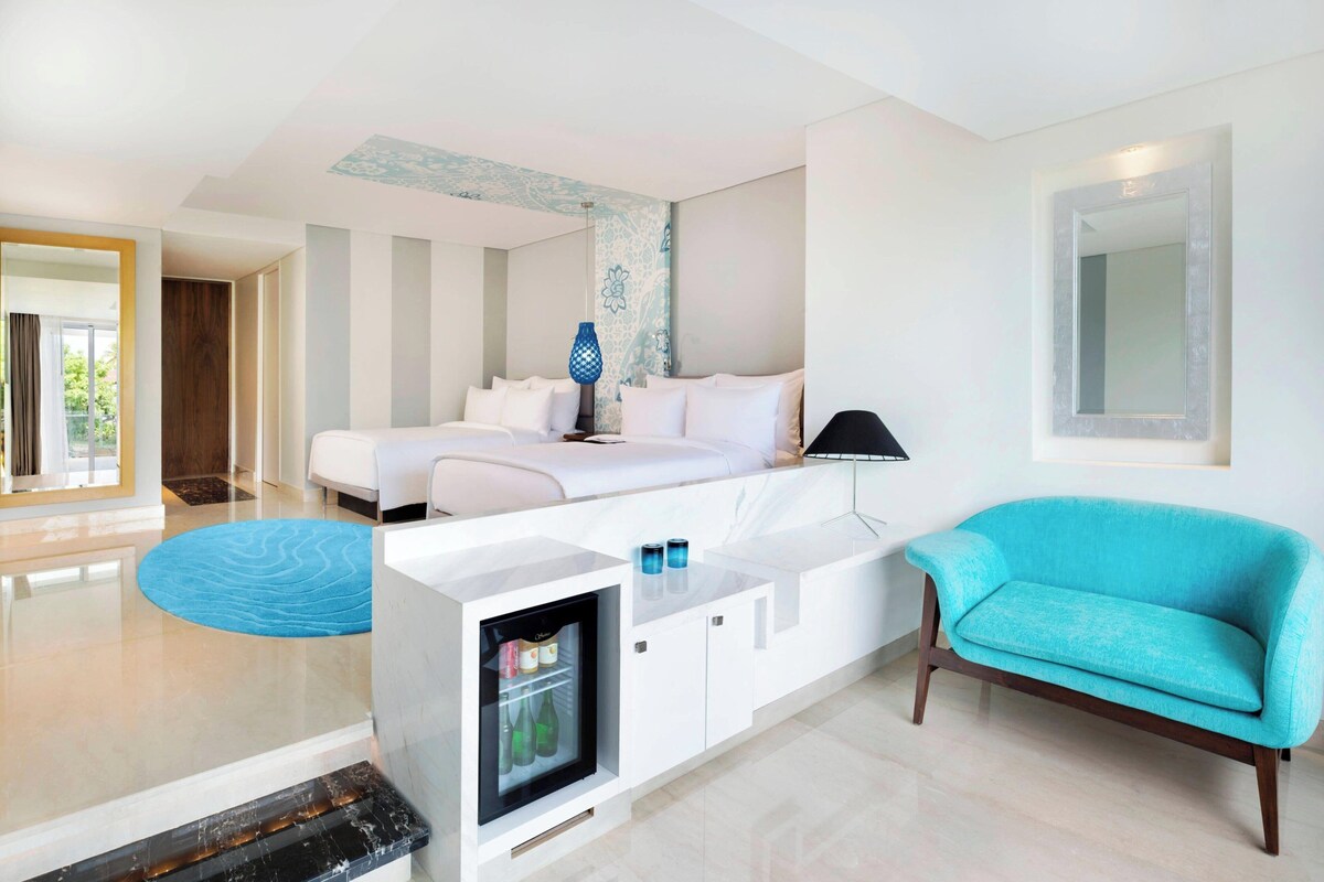 A modern hotel room with a bed, a sofa, a minibar, and a view of the outdoors.