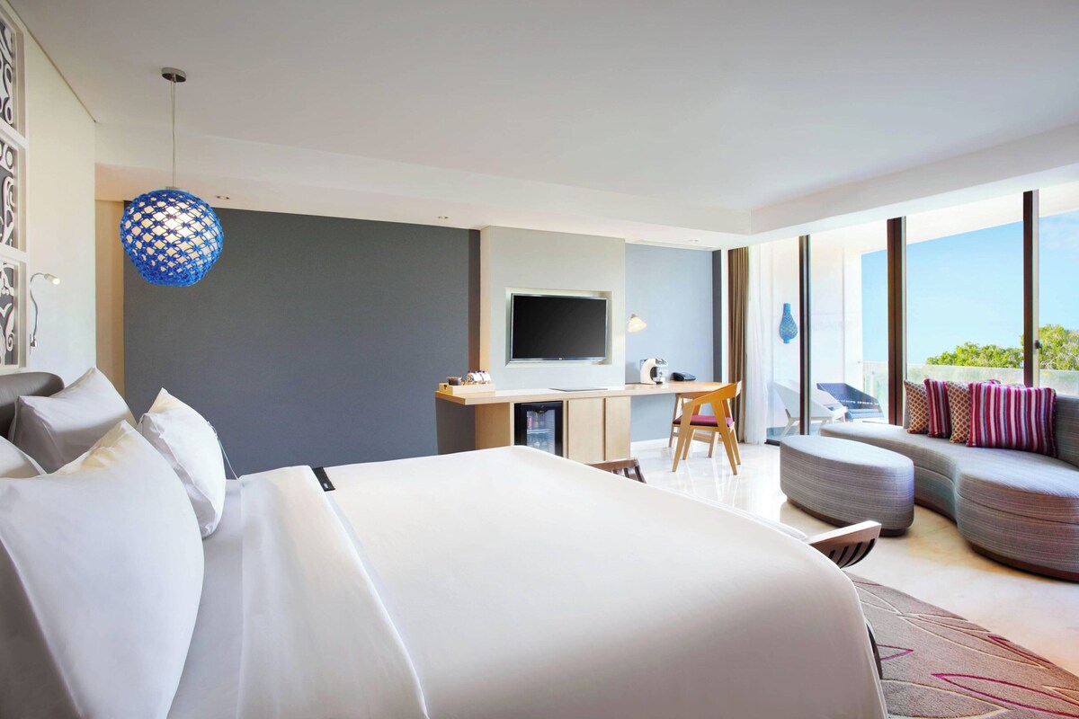 A modern hotel room with a large bed, a desk, a television, and a balcony with seating.