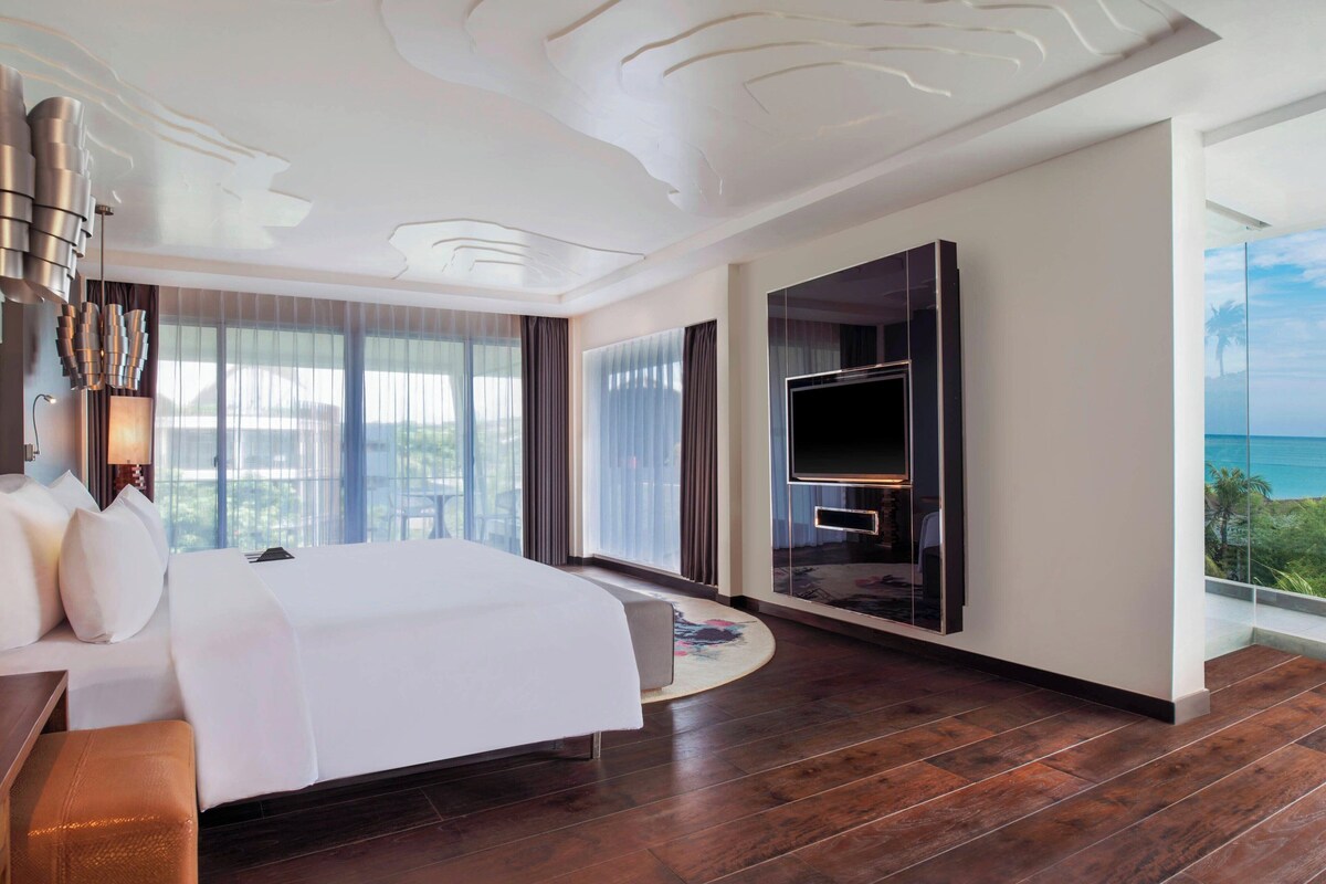 A modern hotel room with a large bed, a flat-screen TV, and a balcony with a view of the ocean.