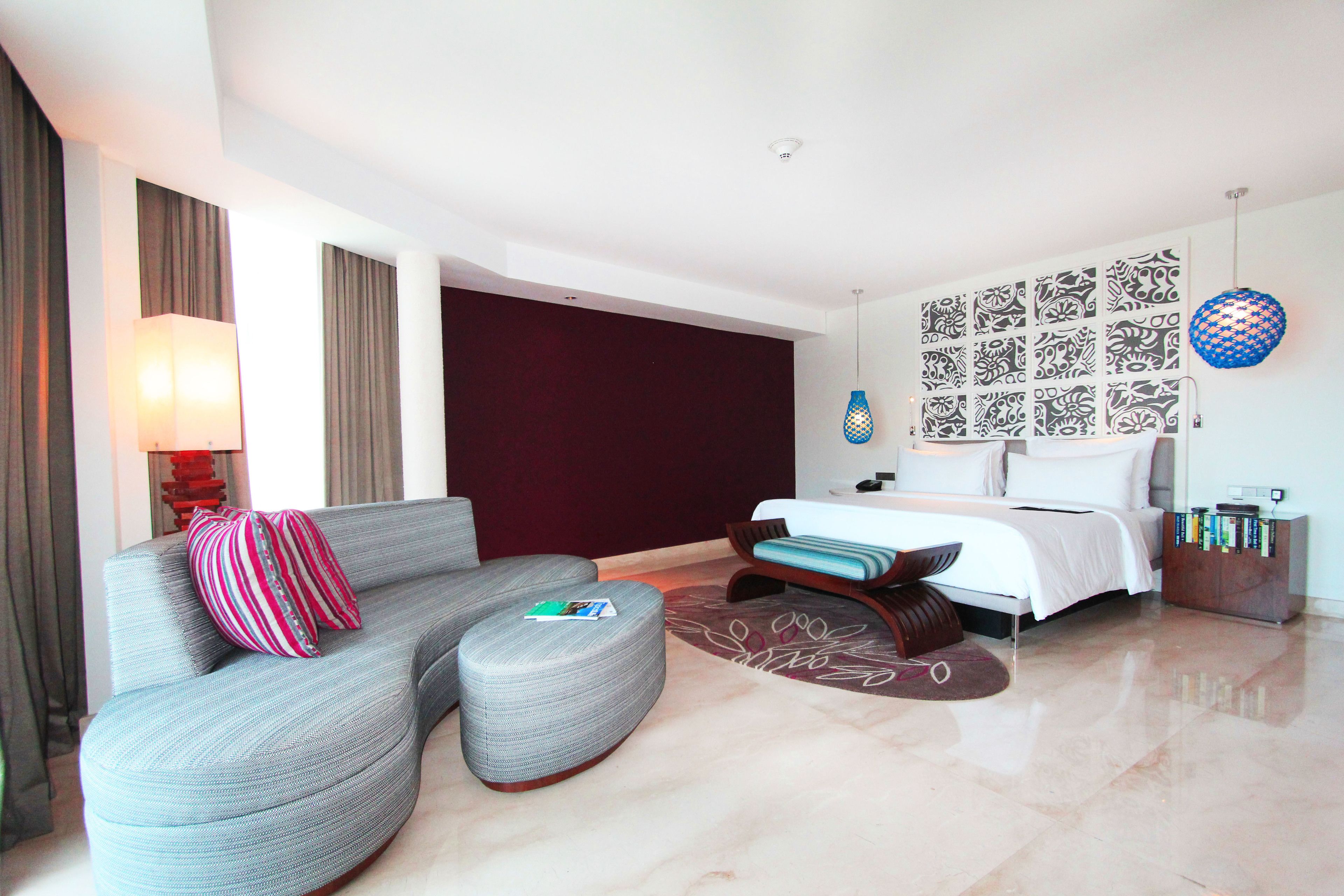 Une chambre d’hôtel moderne avec un canapé gris aux lignes courbes, un lit recouvert de draps blancs, un mur à motifs et un luminaire bleu suspendu.