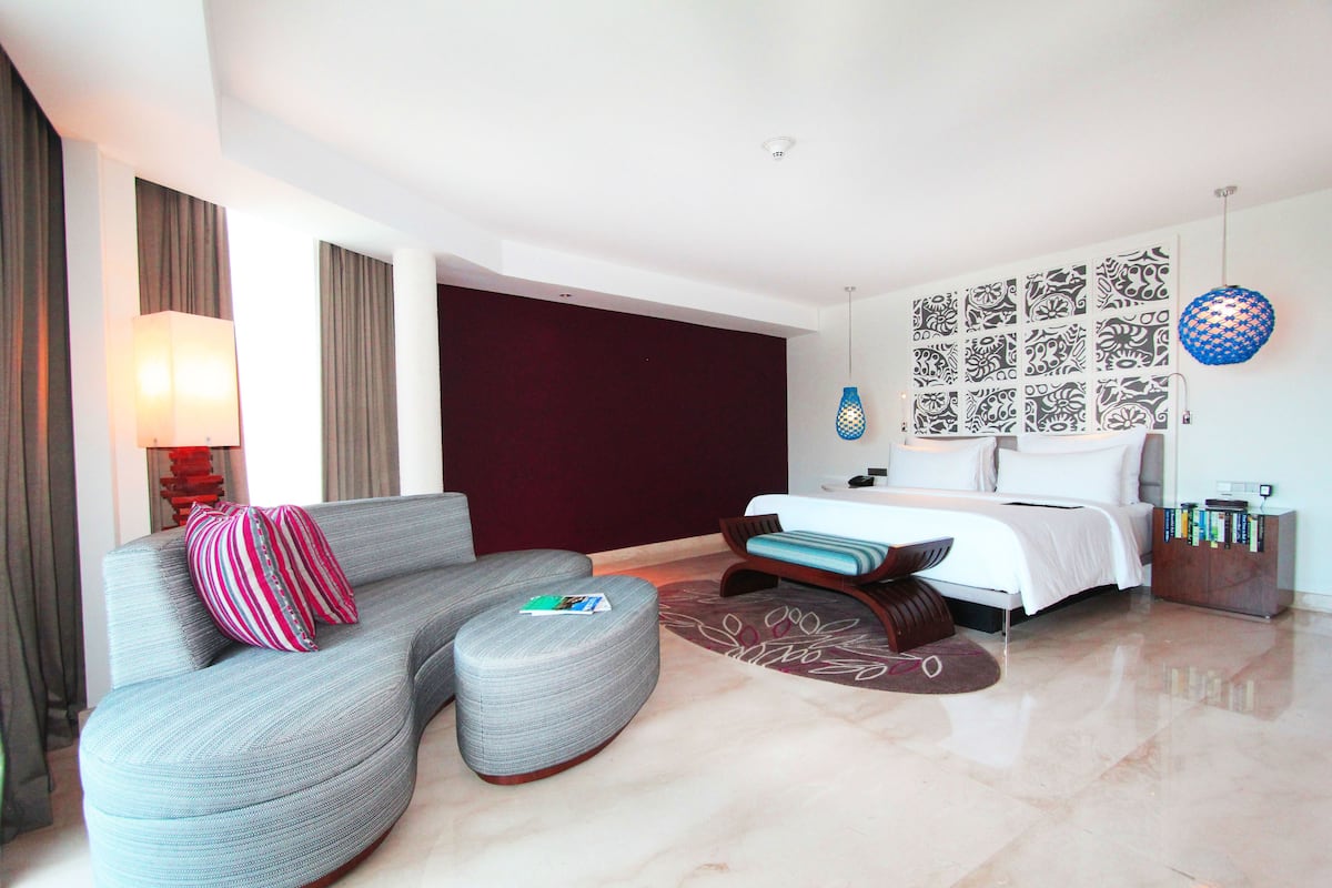 Une chambre d’hôtel moderne avec un canapé gris aux lignes courbes, un lit recouvert de draps blancs, un mur à motifs et un luminaire bleu suspendu.