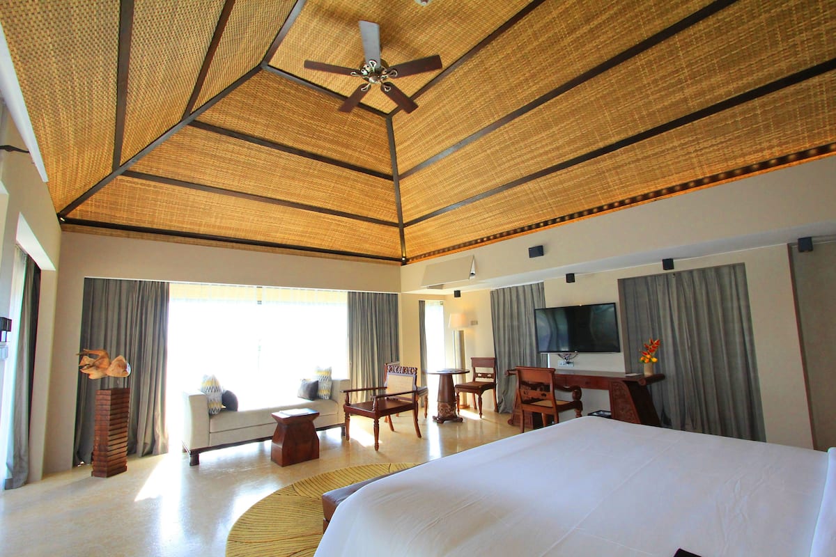 Une chambre d’hôtel spacieuse, dotée d’un grand lit, d’un ventilateur de plafond, d’une télévision à écran plat et d’un coin salon avec un canapé et des fauteuils.