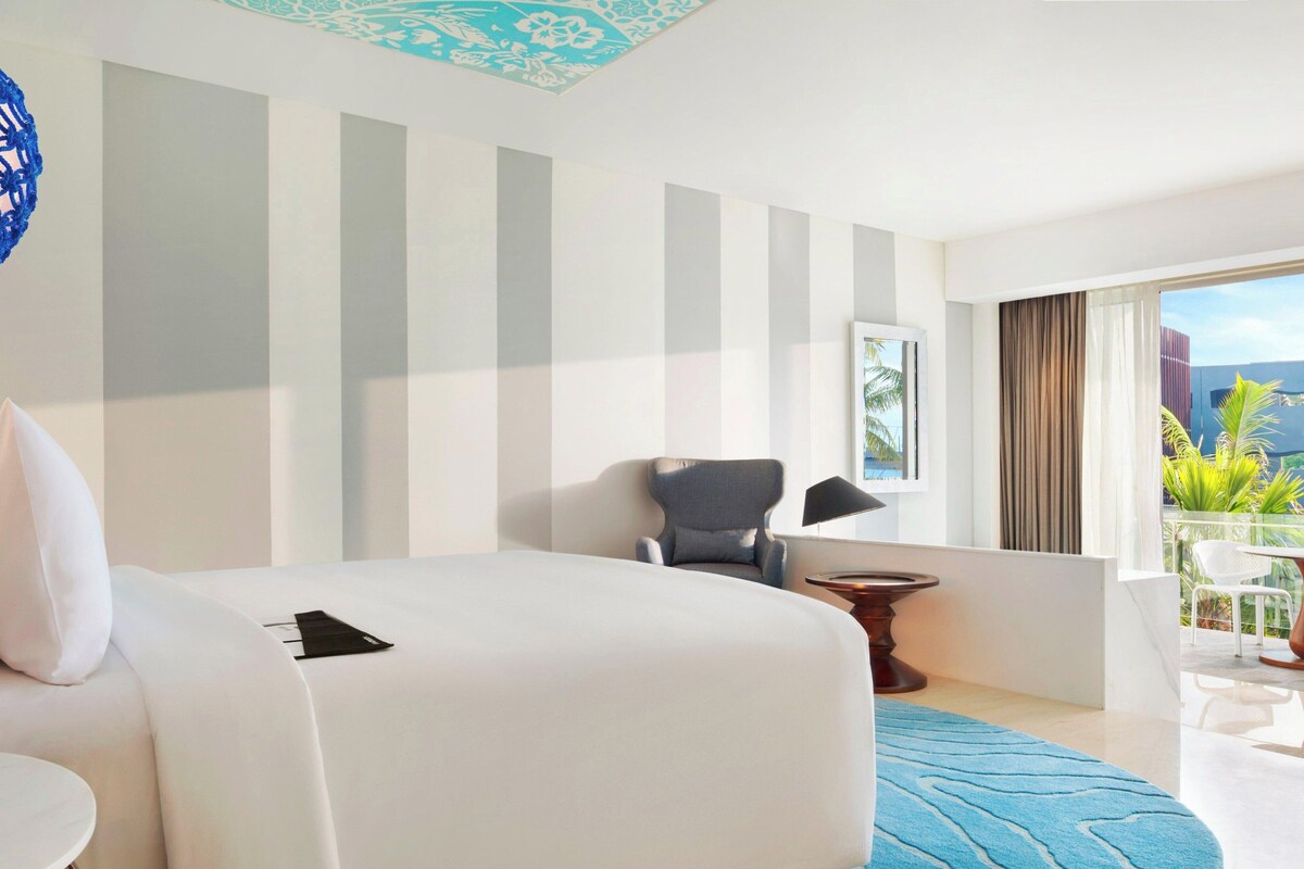 A hotel room with a bed, a chair, a small table, a window with a view of greenery, and a patterned ceiling.
