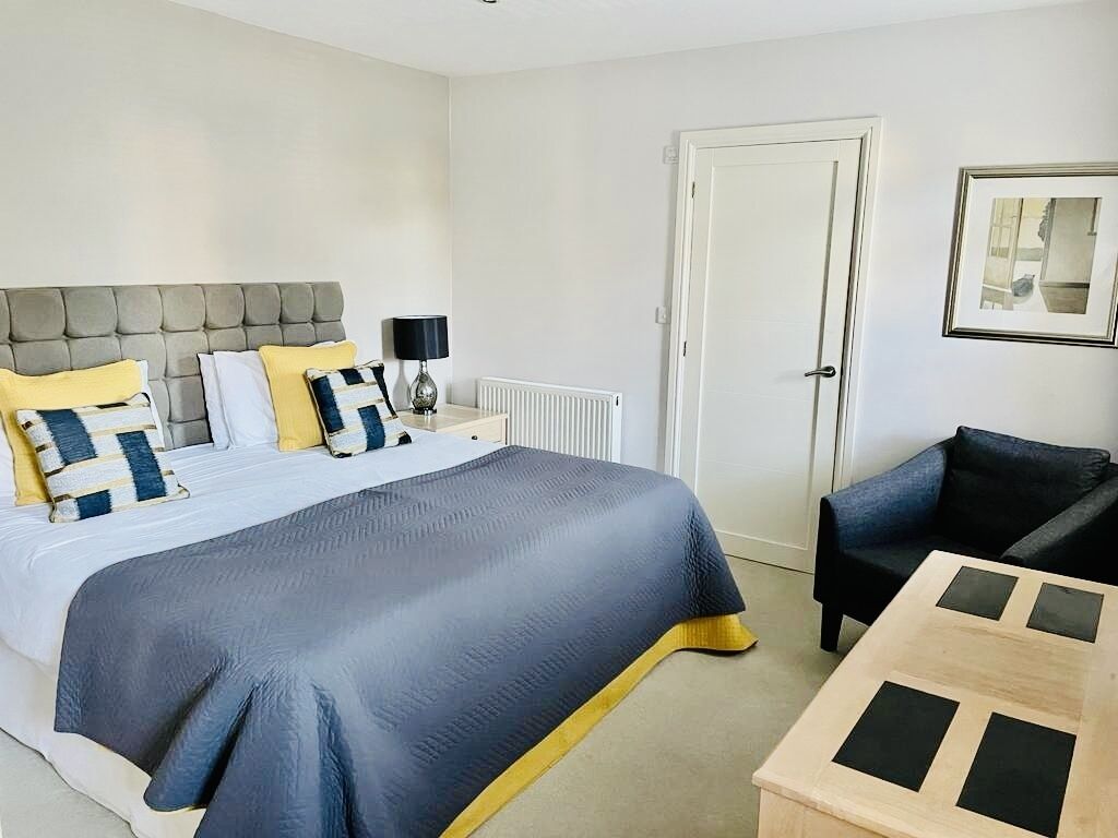 A hotel room with a large bed, a bedside table with a lamp, a chair, and a framed picture on the wall.