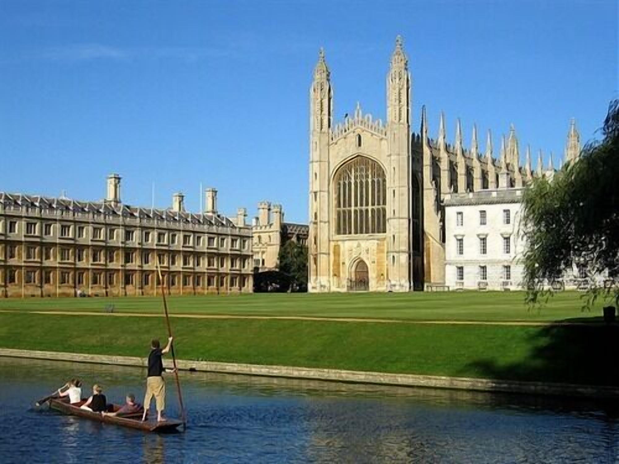 A historic building with a large arched entrance and tall spires, a river with a rowing boat, and a well-maintained lawn.