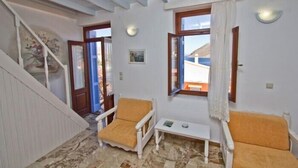 Una habitación con una escalera, dos sillas blancas con cojines amarillos, una mesita y un ventanal con vistas al mar.