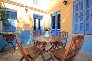 A round wooden table with six chairs, surrounded by blue shutters and a potted plant. - Dorian Hotel (Symi)