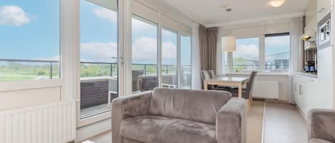 A modern living room with a sofa, a dining table, and chairs. There is a kitchen area with a microwave and cabinets. Large windows provide a view of the outdoors.