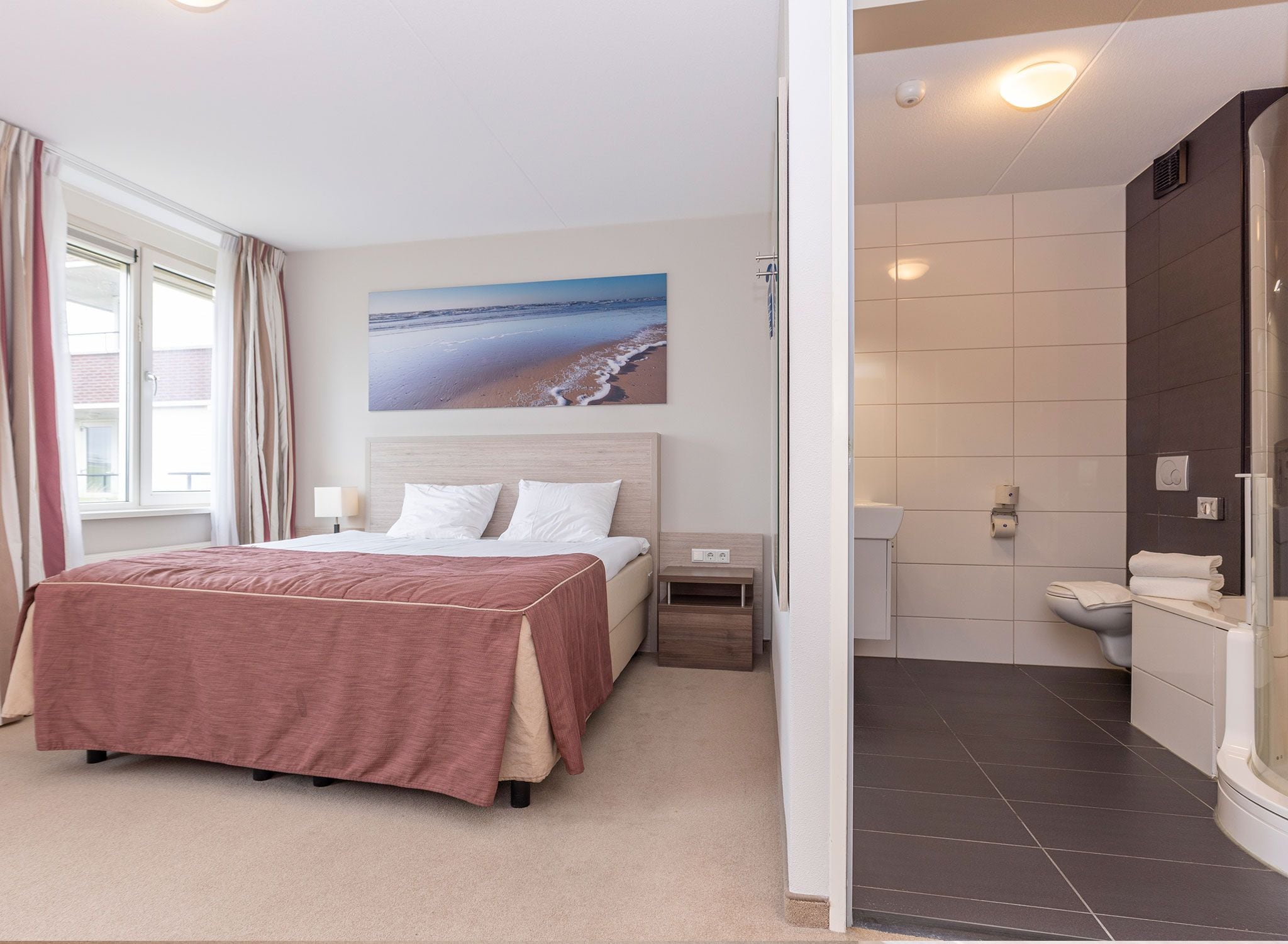Ein Schlafzimmer mit einem Bett, einem Nachttisch, einem Fenster mit Vorhängen und einem Wandbild mit Strandmotiv.
