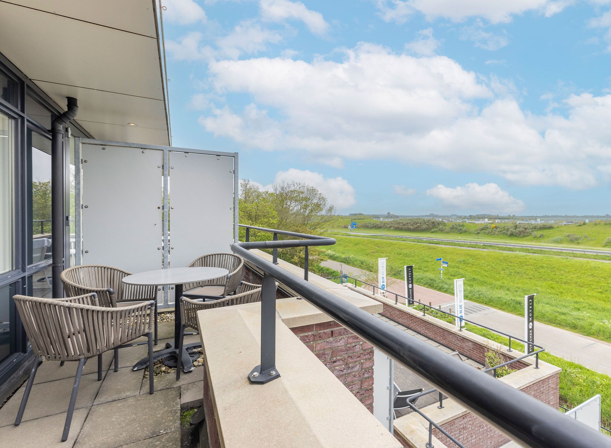 A balcony with outdoor seating, a view of a green landscape, and a clear sky.