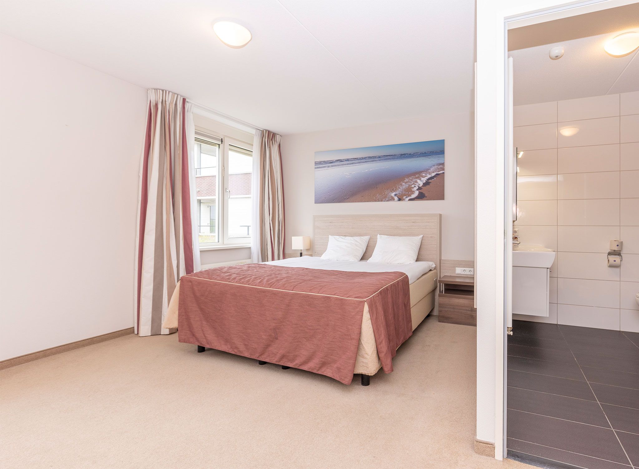 Une chambre à coucher avec un lit, une fenêtre avec des rideaux, un tableau représentant une plage, et une salle de bain avec des toilettes et un lavabo.