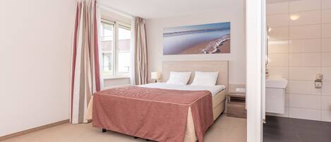 Un dormitorio con cama, una ventana con cortinas, un cuadro de una playa y un baño con inodoro y lavabo.