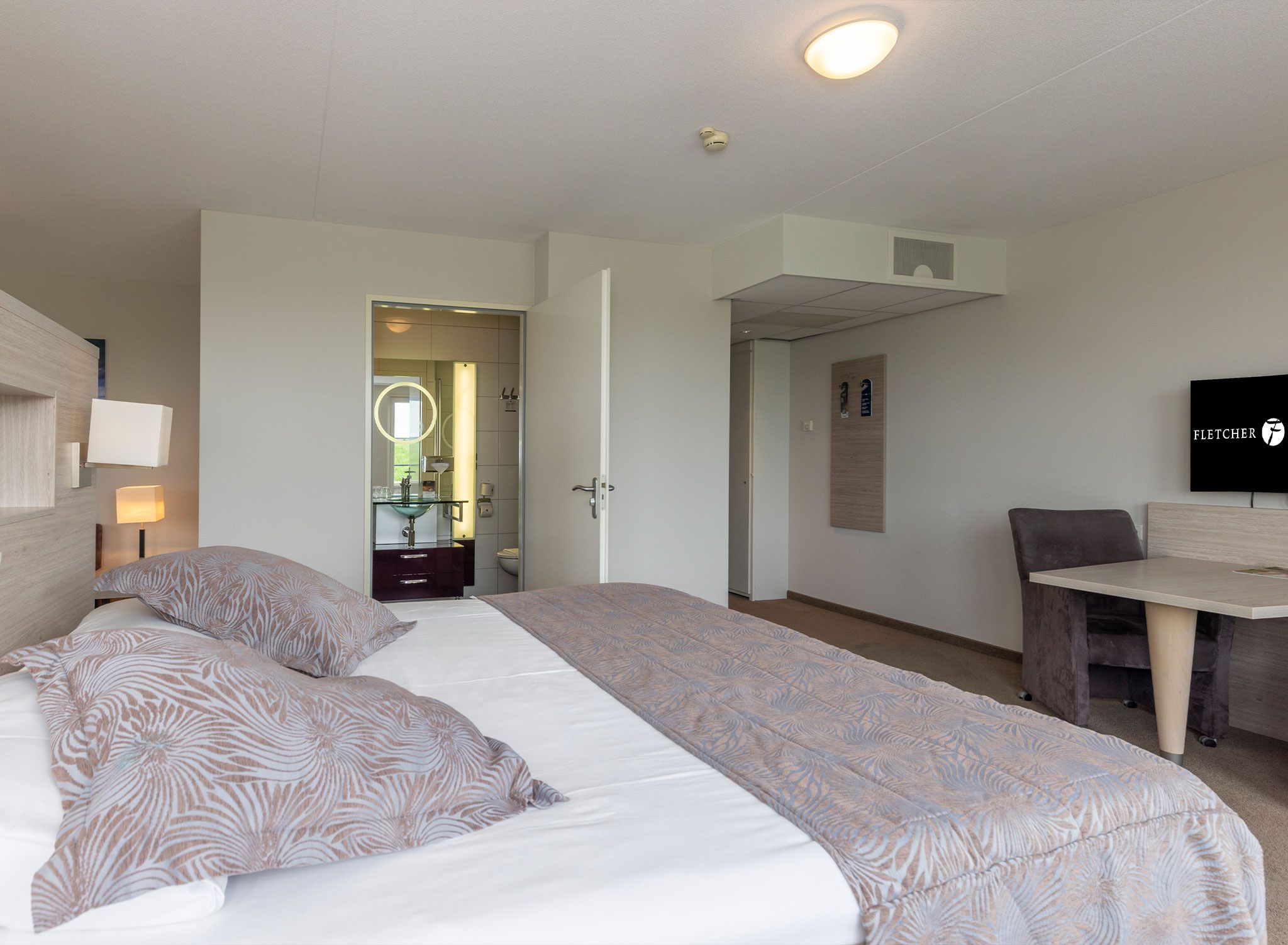 Habitación de hotel con cama grande, escritorio, silla, televisor y baño visible a través de una puerta abierta.