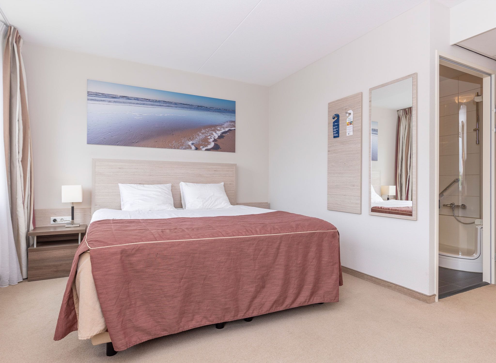 Habitación de hotel con cama, mesita de noche, lámpara, un cuadro de playa, espejo y zona de ducha.
