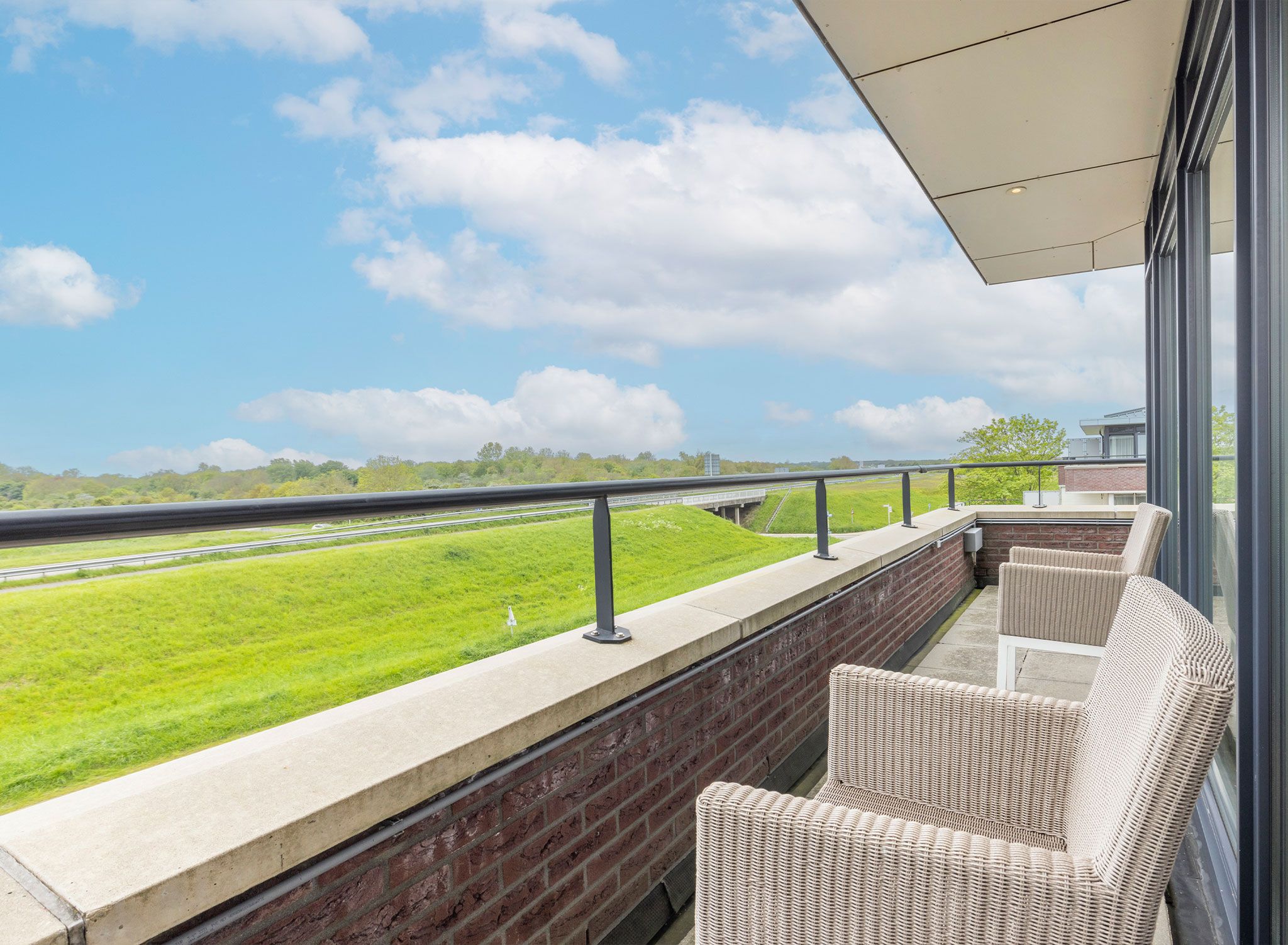 A balcony with wicker furniture overlooking a green landscape and a highway.