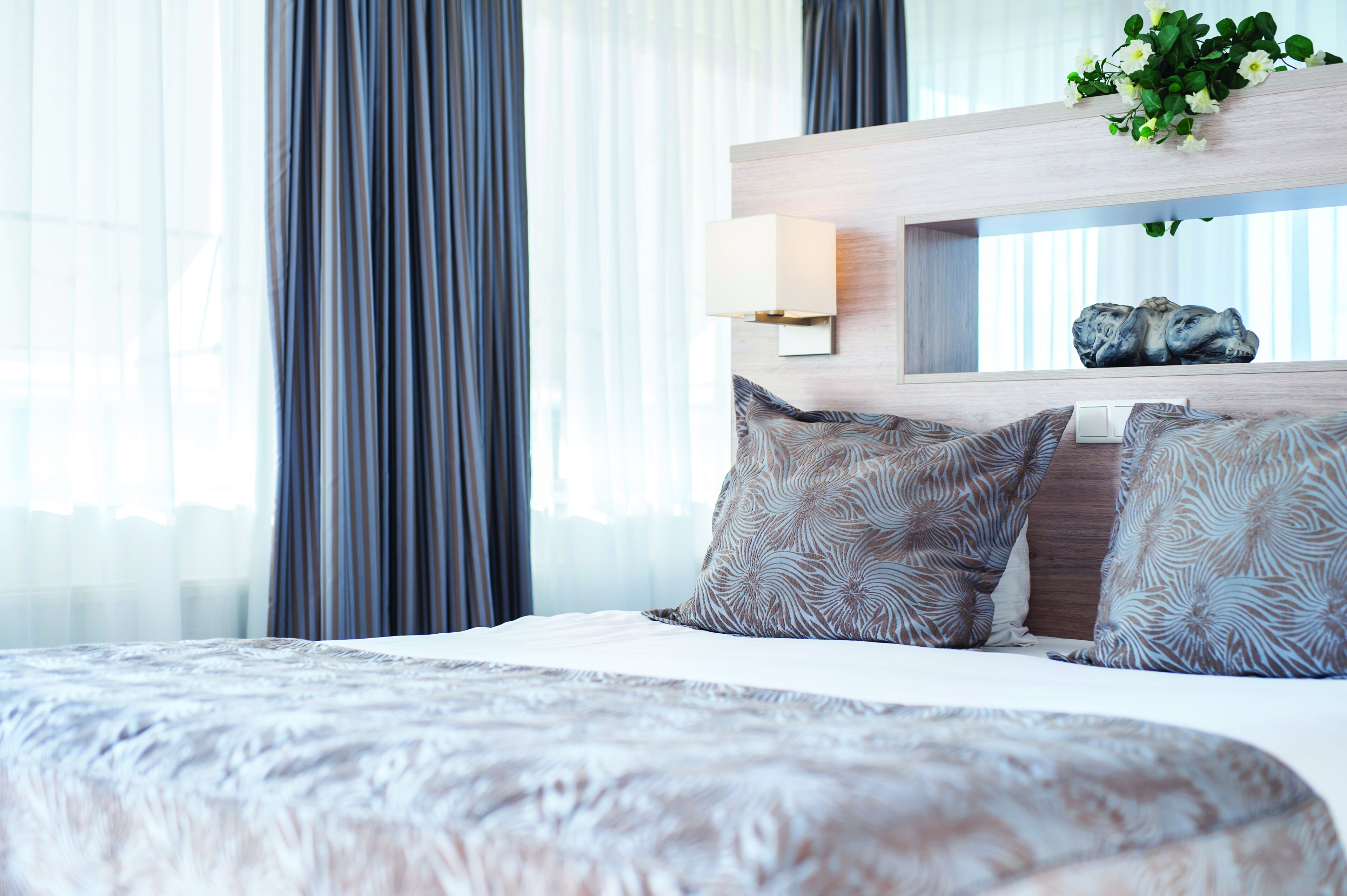 Una habitación de hotel moderna con una cama grande, una mesita de noche con lámpara y una planta decorativa en la cabecera.