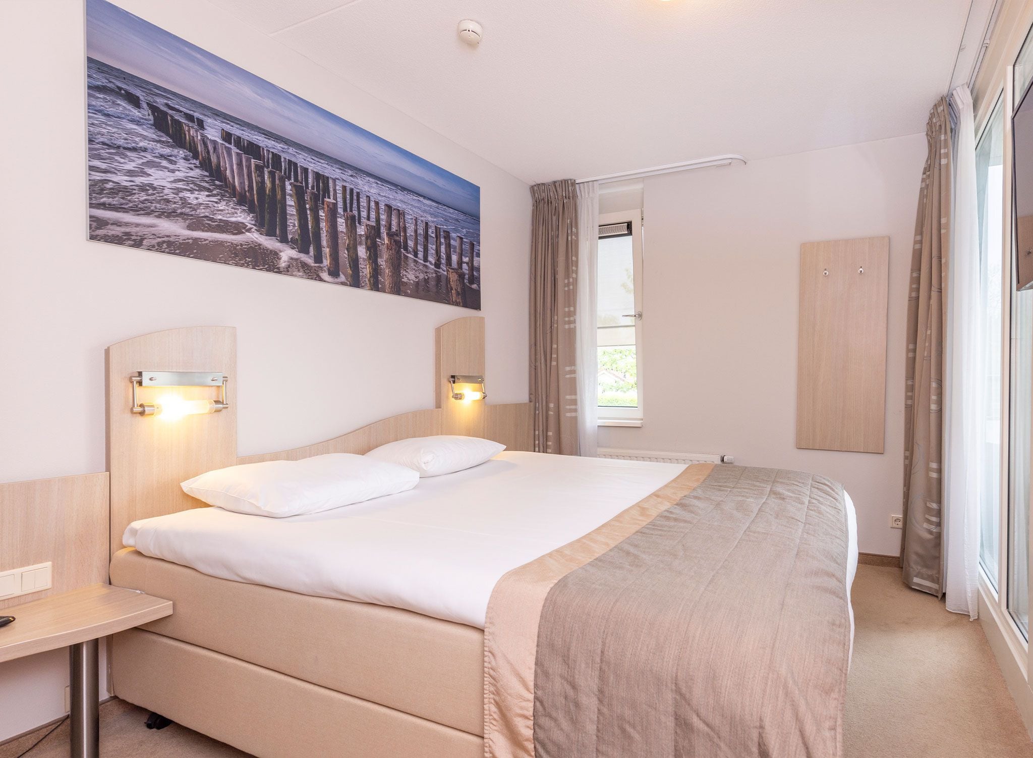 A hotel room with a large bed, bedside tables, a painting on the wall, and a window with curtains.