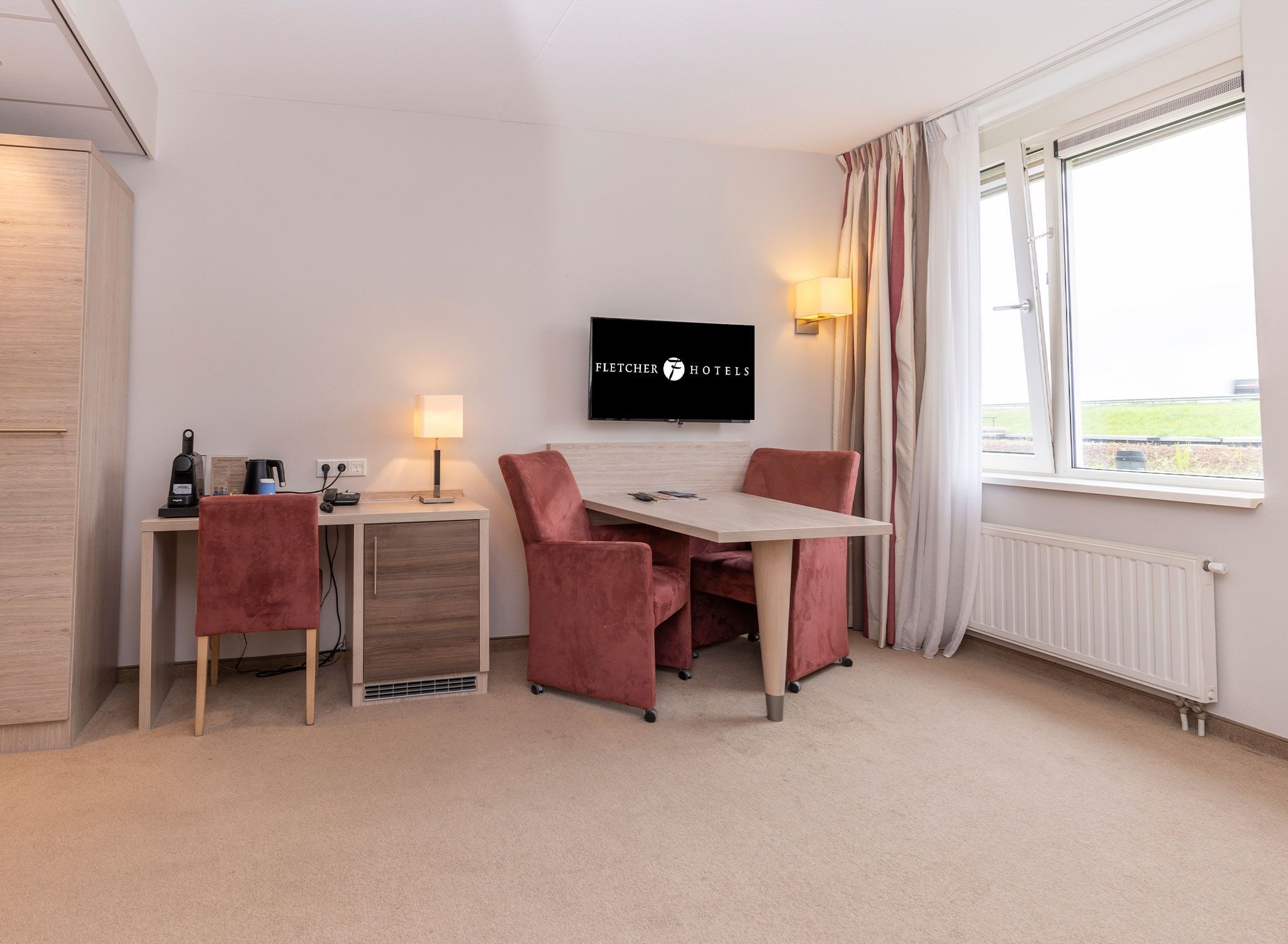 A hotel room with a desk, two chairs, a television, a lamp, and a window with curtains.