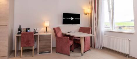 Habitación de hotel con escritorio, dos sillas, televisor, lámpara y ventana con cortinas.