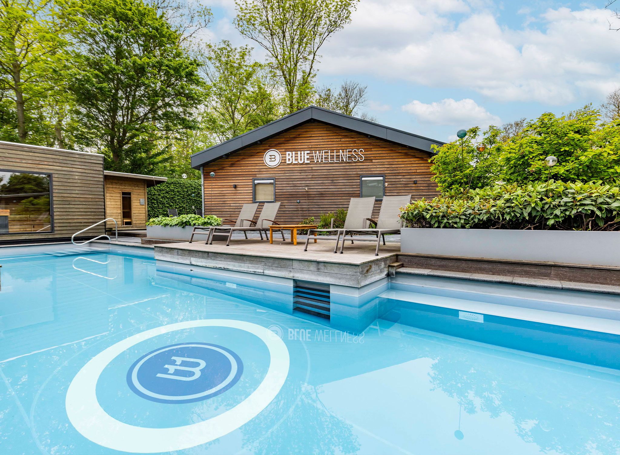 A modern pool area with lounge chairs, a wooden building labeled 'BLUE WELLNESS,' and a clear blue pool.