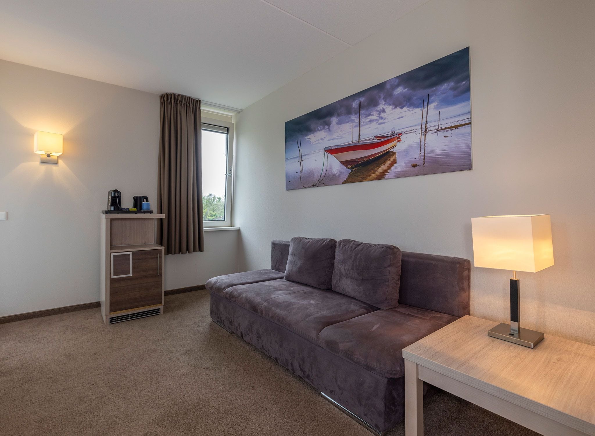 A room with a sofa, a table with a lamp, a painting of boats on water, a window with curtains, and a shelf with items.