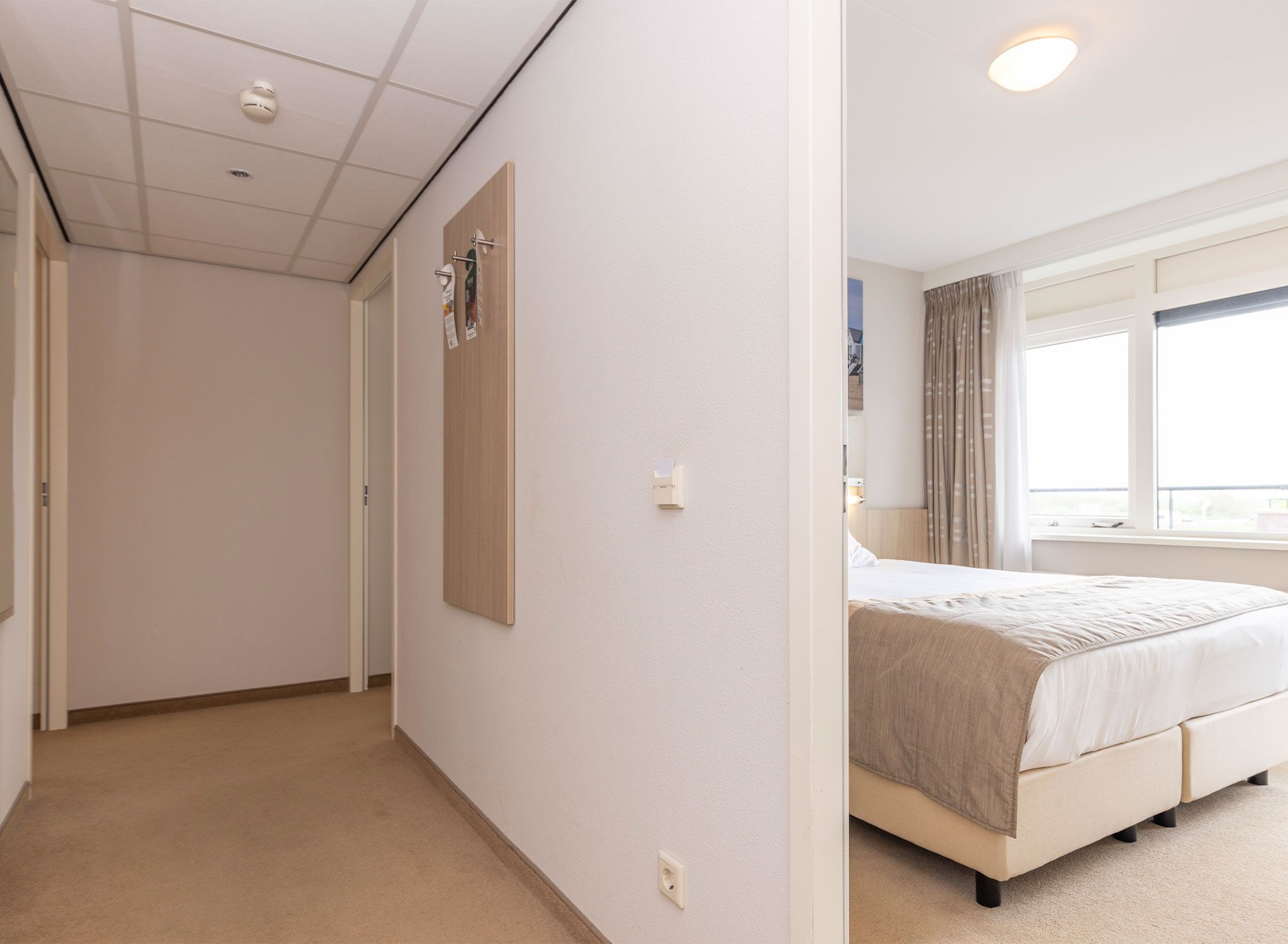 Une chambre d’hôtel avec un lit, une fenêtre avec des rideaux, une porte et un luminaire fixé au mur.