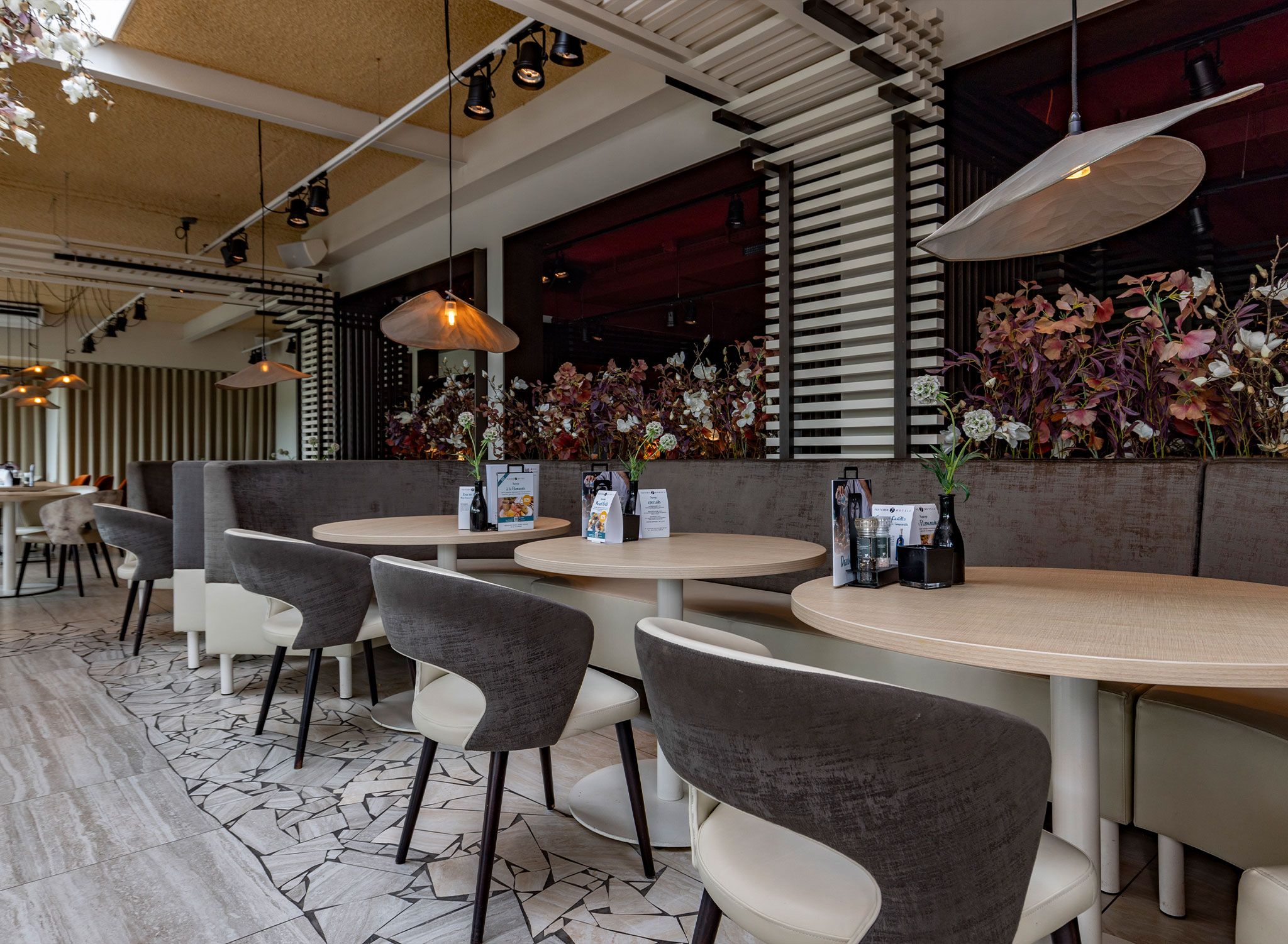 A restaurant interior with round tables, grey and white chairs, hanging pendant lights, and a wall adorned with floral arrangements.