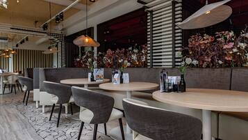 A restaurant interior with round tables, grey and white chairs, hanging pendant lights, and a wall adorned with floral arrangements.
