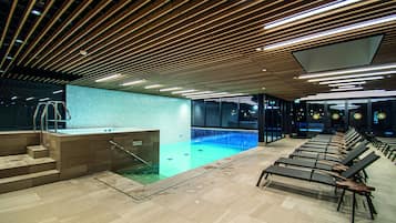 Ein moderner, überdachter Poolraum mit Holzbalkendecke, einer kleinen Poolanlage mit Stufen und Liegestühlen entlang des Beckens.