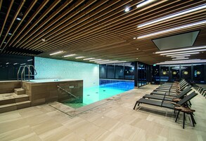 A modern indoor swimming pool with a wooden ceiling, a small pool area with steps, and lounge chairs along the poolside.