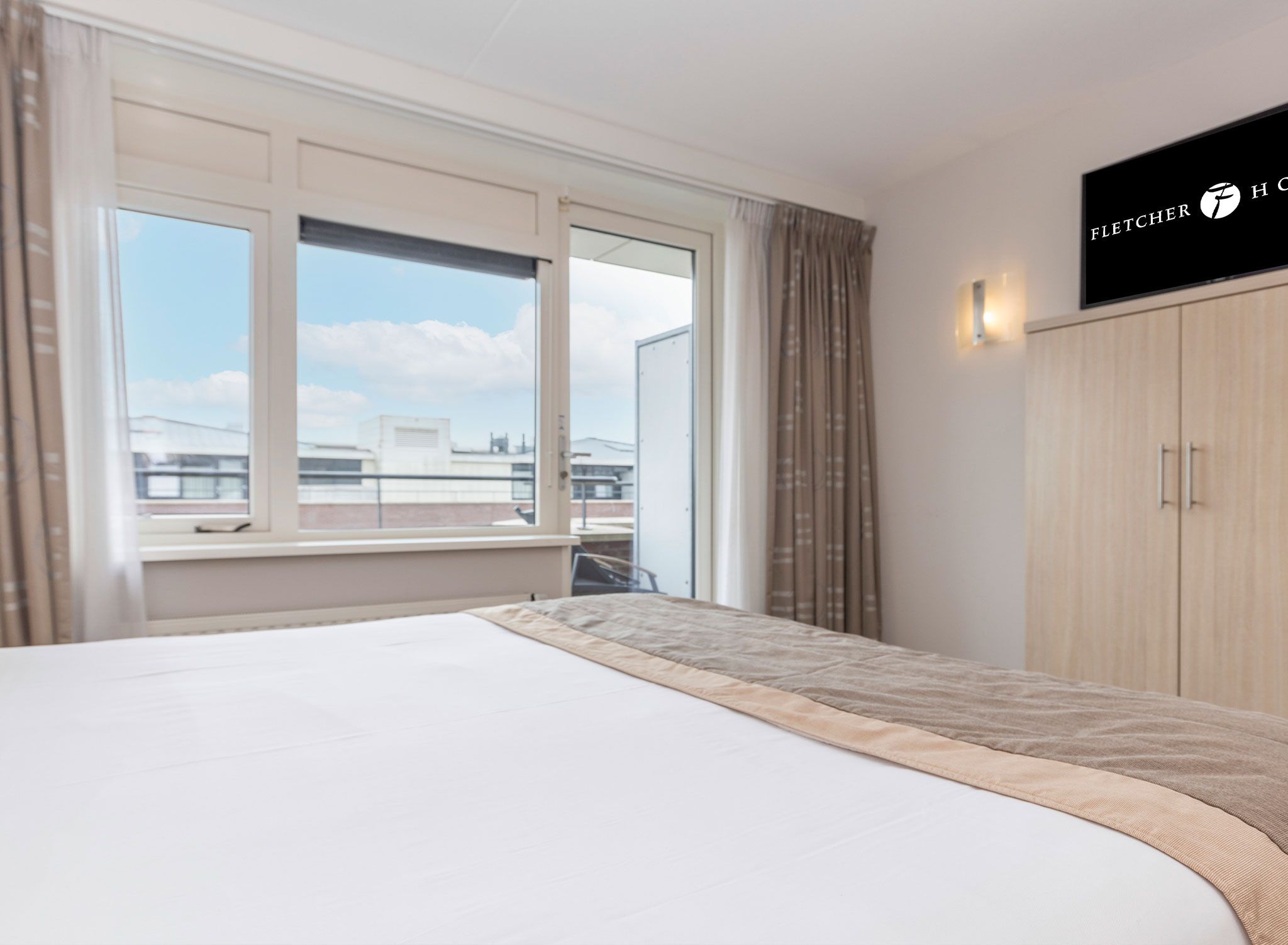 A hotel room with a large bed, a window with curtains, a wardrobe, and a television on the wall.