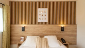 Habitación de hotel con cabecera de madera, dos mesitas de noche, cama con ropa blanca, un cuadro enmarcado y un teléfono sobre una de las mesitas.