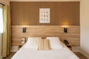 Habitación de hotel con cabecera de madera, dos mesitas de noche, cama con ropa blanca, un cuadro enmarcado y un teléfono sobre una de las mesitas.