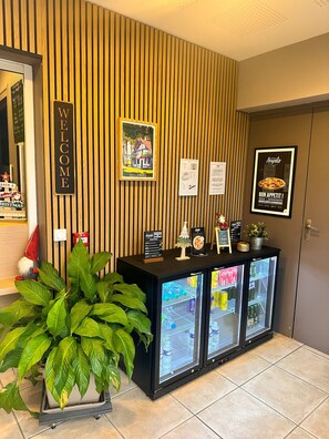 Zona de recepción con un cartel de 'WELCOME', un cartel enmarcado que dice 'LES LANDES', un tablero de menú y un refrigerador con bebidas.