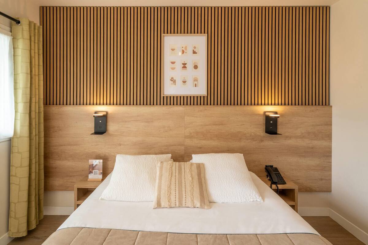 A hotel room with a wooden headboard, two bedside lamps, a framed artwork, a bedside phone, and a bed with white bedding.