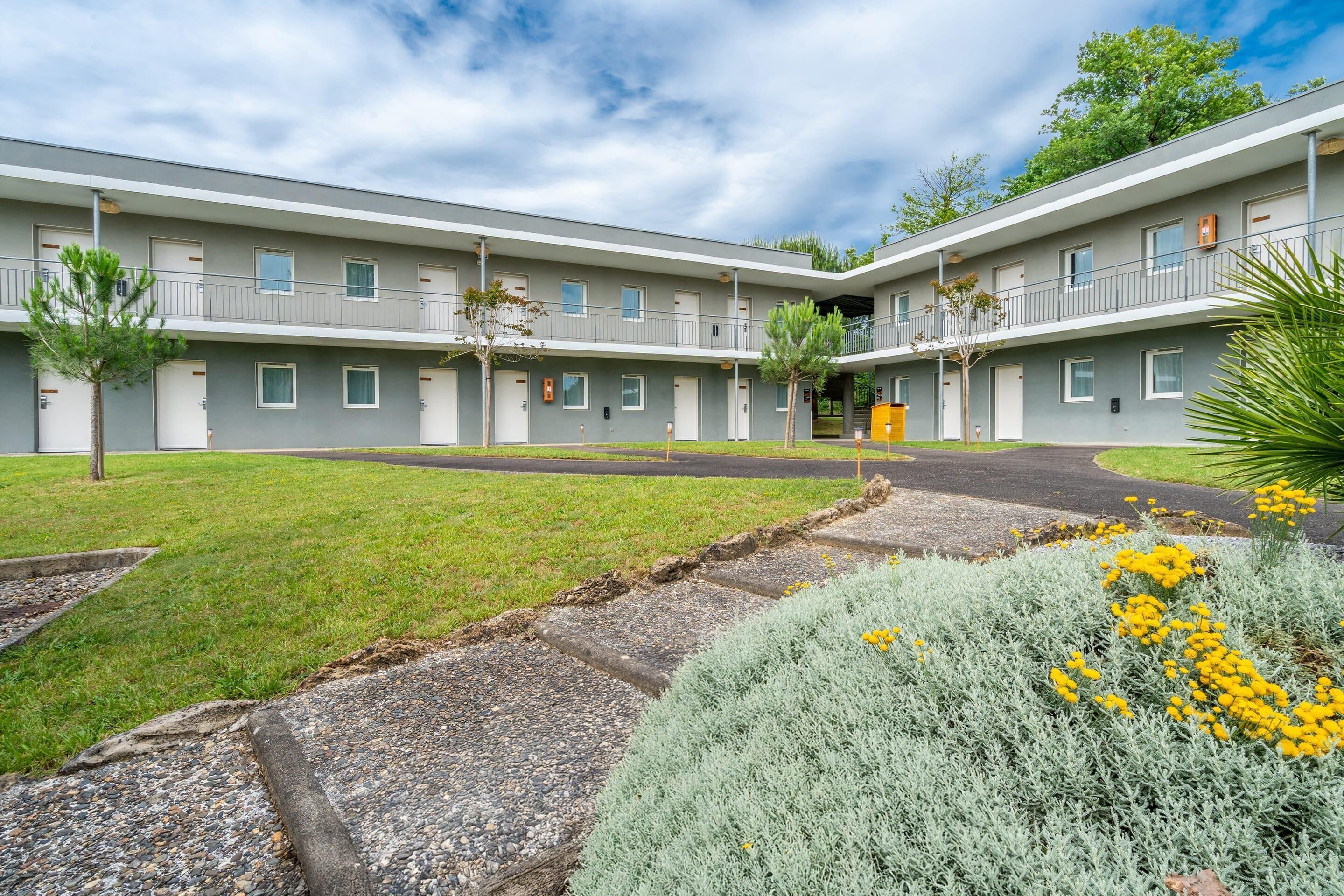 A multi-story motel with a central courtyard, featuring a grassy area, a paved walkway, and a variety of plants.