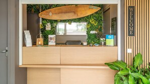 Un mostrador de recepción con una decoración de tabla de surf, un cartel que dice 'WELCOME' y una pared verde con plantas.