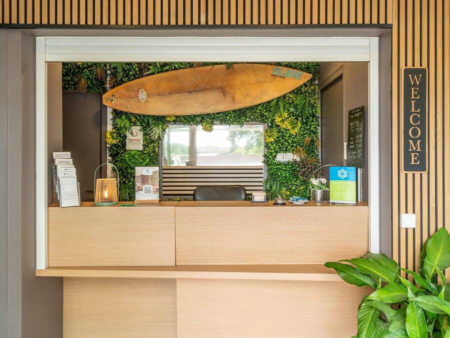 Un comptoir d’accueil décoré avec une planche de surf, une pancarte « BIENVENUE » et un mur végétal.