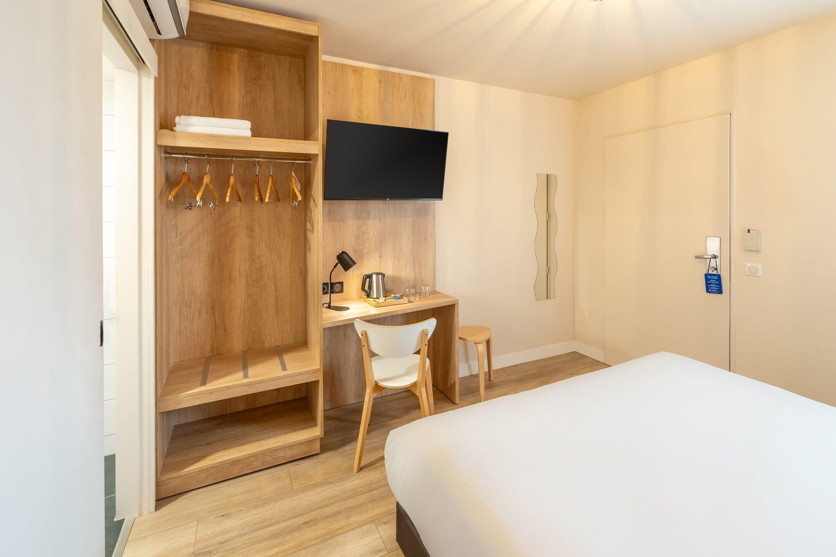 Habitación de hotel con cama, escritorio con silla, televisor montado en la pared y armario con ropa colgada.
